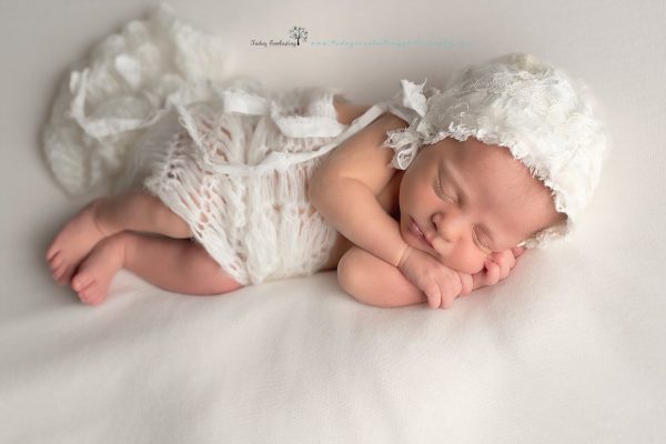 Sleeping baby girl wearing white lacy bonnet, with white lacy wrap on soft white background