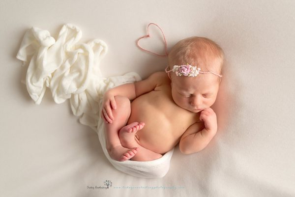 Newborn baby girl wearing pink rosebud headband with ends shaped into a heart, with white wrap, on soft white background