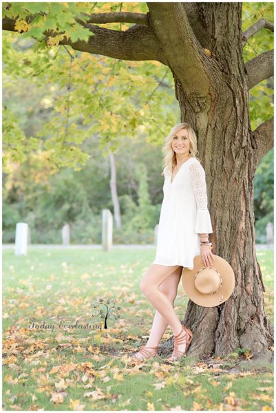 Beautiful high school graduate in white lacy dress leaning against large tree in hazy golden light