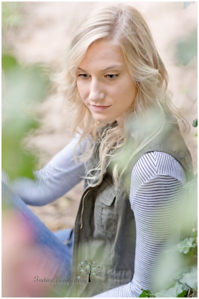 Sweet young lady with soft blonde curls and a gentle smile seen through soft green foliage.