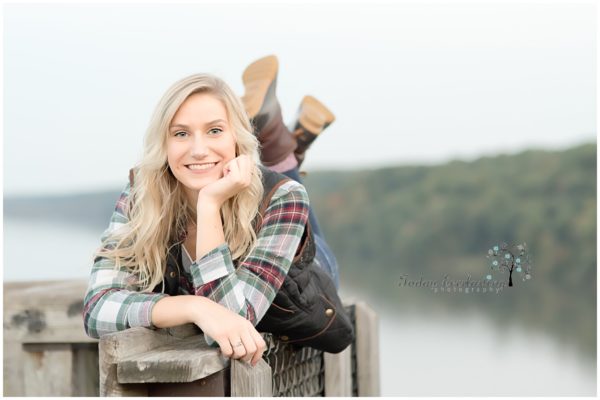 Lovely blonde high school student with long hair and plaid flannel shirt, perched on a railing with her feet kicked up behind her against a stunning scenic river background.