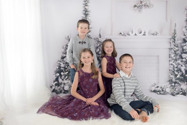 Children wearing holiday outfits seated in front of white mantel