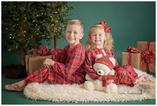 A boy and a girl dressed in plaid Christmas pajamas sitting on a cream blanket
