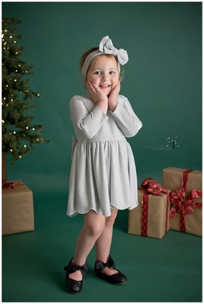 A little girl wearing a silver dress with a matching headband standing on a dark green backdrop with Christmas decorations