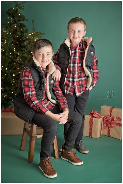 Two boys in black pants and vests with plaid shirts posed on a dark green background with Christmas decorations