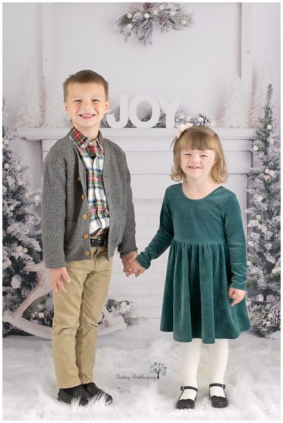 A boy and a girl dressed in Christmas outfits standing in front of a white mantel decorated with JOY.