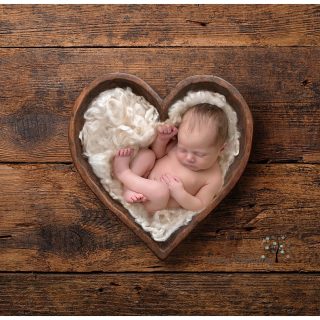 A newborn curled up on curly wool in a wooden bowl shaped like a heart