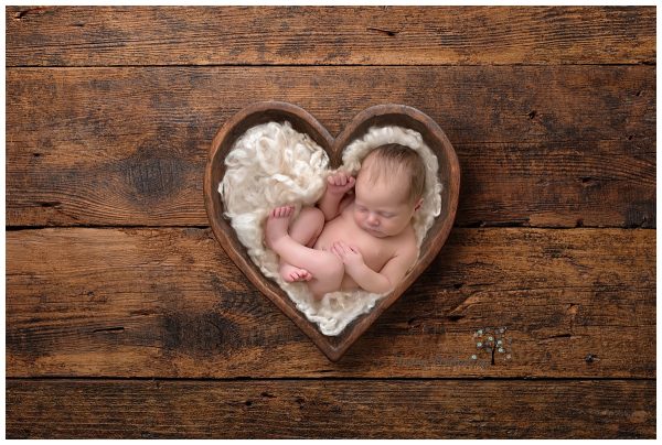A newborn curled up on curly wool in a wooden bowl shaped like a heart