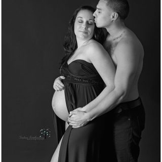 Black & White studio maternity portrait of a couple. The woman is wearing a strapless black gown, and her husband is gently kissing the side of her face.