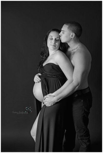 Black & White studio maternity portrait of a couple. The woman is wearing a strapless black gown, and her husband is gently kissing the side of her face.