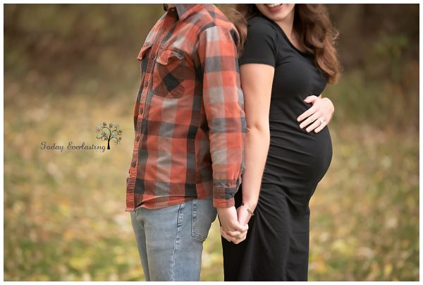 Outdoor portrait of a couple standing back to back holding hands. The woman is pregnant and has a hand resting on the top of her baby bump.