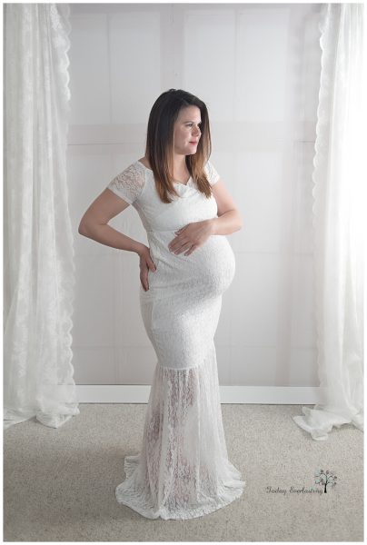 Pregnant woman wearing a white gown, with a white background. She has one hand on her baby bump, and the other hand is resting on her hip.