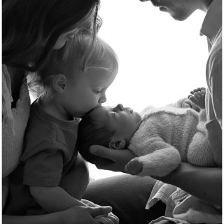 Toddler kissing newborn baby while parents look on during a newborn portrait session in Morris IL